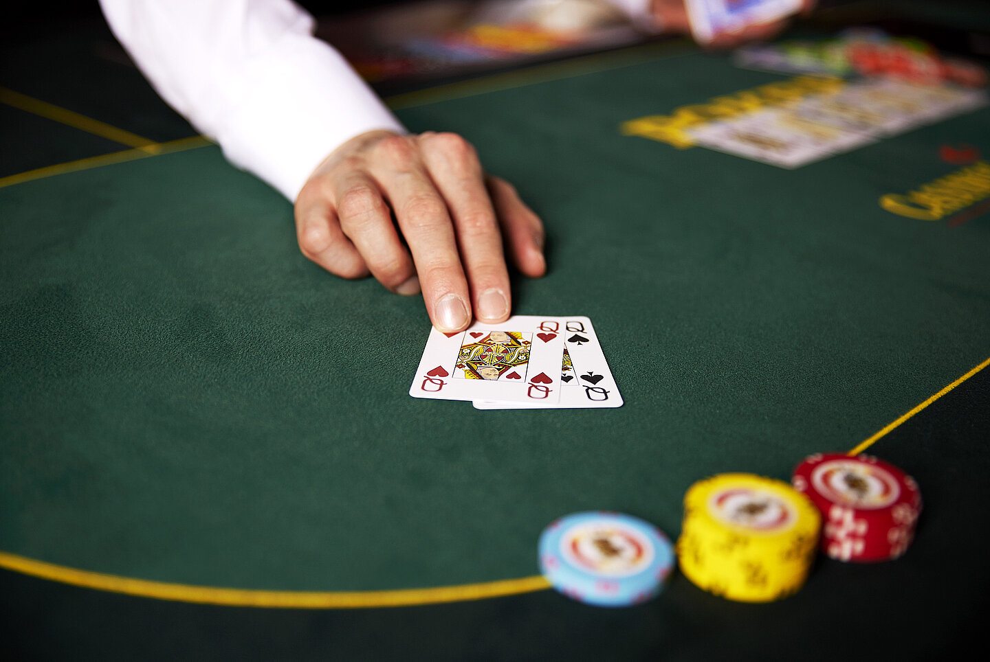 Hand von Croupier schiebt zwei Damen Poker Karten über den grünen Tisch, daneben bunte gestapelte Jetons und verschiedene Karten