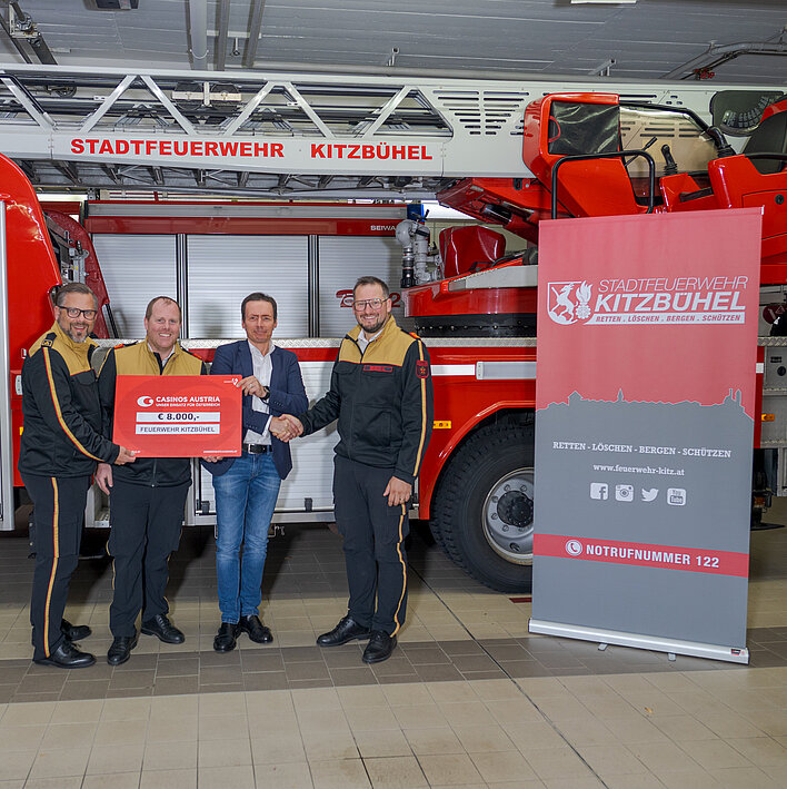 3 Mitglieder der Stadtfeuerwehr Kitzbühel und Casino Kitzbühel Mitarbeiter bei Scheck-Übergabe vor einem Feuerwehr-Wagen
