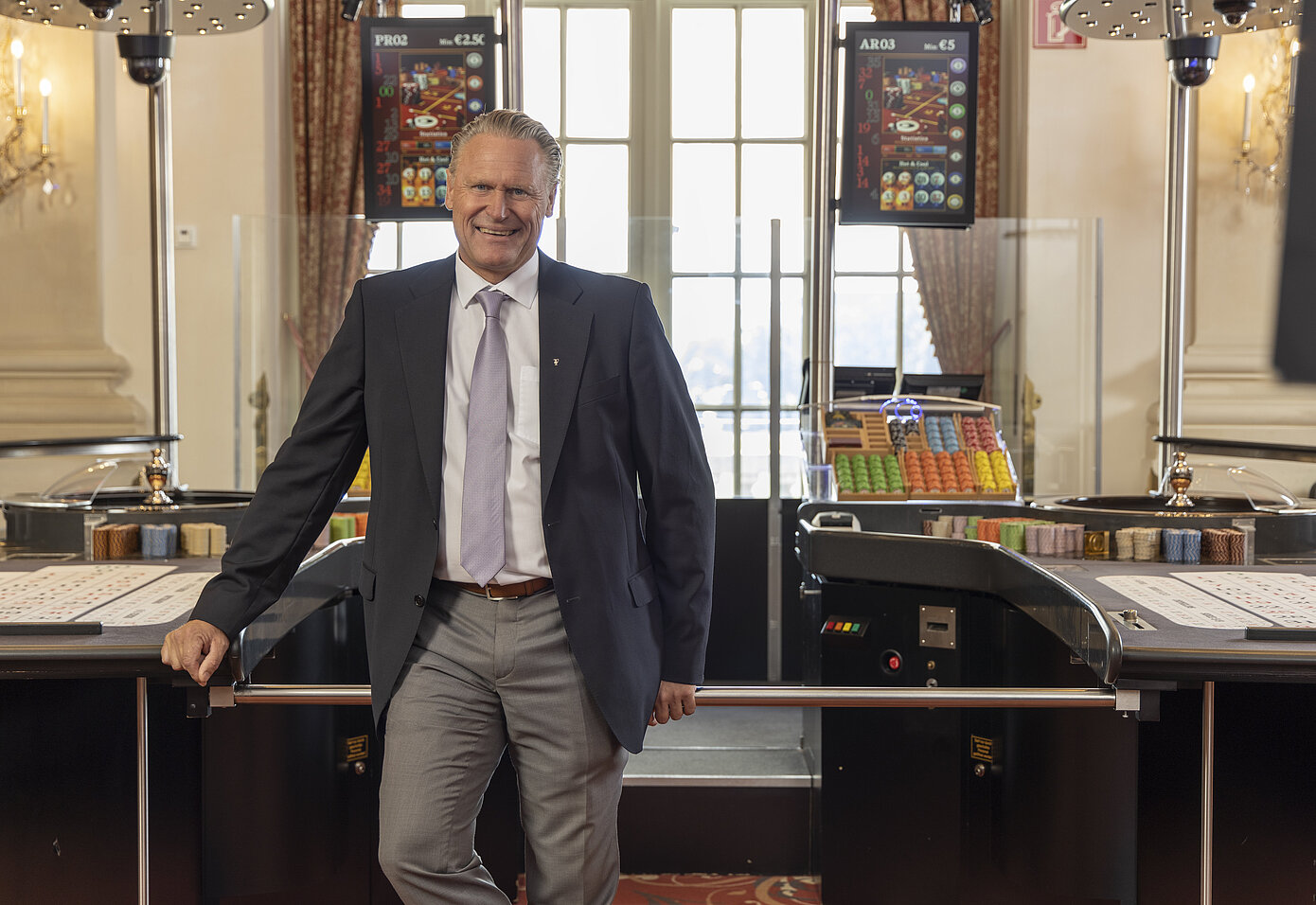 Mann mittleren Alters, helle Haare zurückgekämmt, mit weißem Hemd, blauer Krawatte, grauer Anzugshose und schwarzem Sacko stehend zwischen zwei Spieltischen im Casino Zell am See